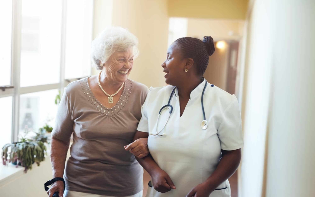 Front view of a caretaker and elderly woman walking and laughing together with linked arms