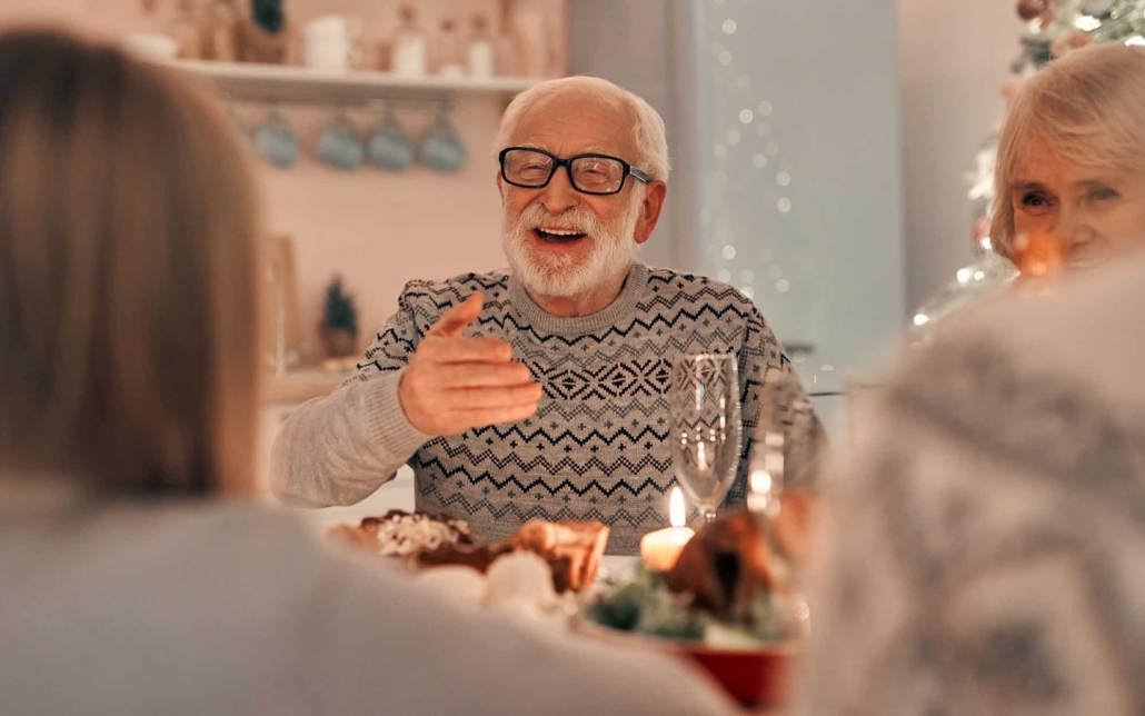 Front View Of A Senior Man Sitting At The Table Surrounded By Family On The Holidays