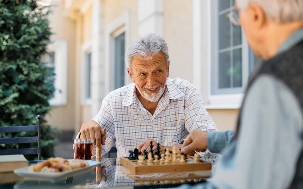 Happy senior man talks to his friend while playing chess on patio at retirement community