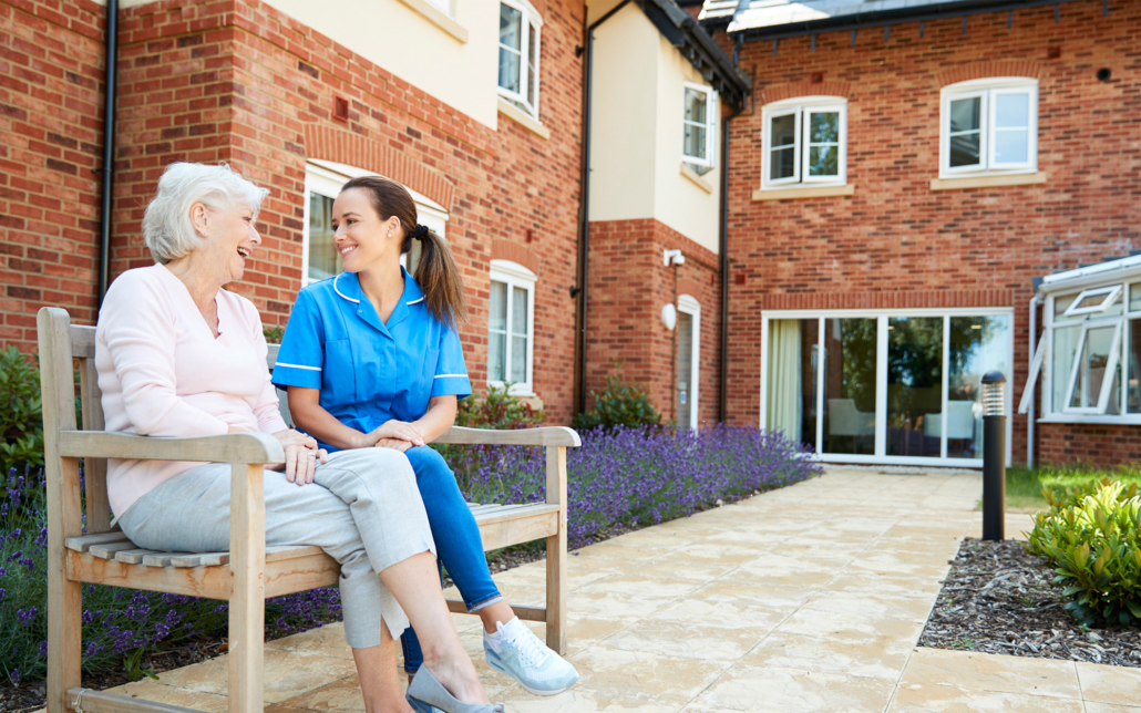 Senior Woman Sitting On Bench And Talking With Nurse In Retirement Home